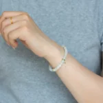 A person in a light gray shirt showcases the Light Green and White Jadeite Bracelet—carved beads with gold-tone spacer details—on their wrist, paired with a simple gold ring.