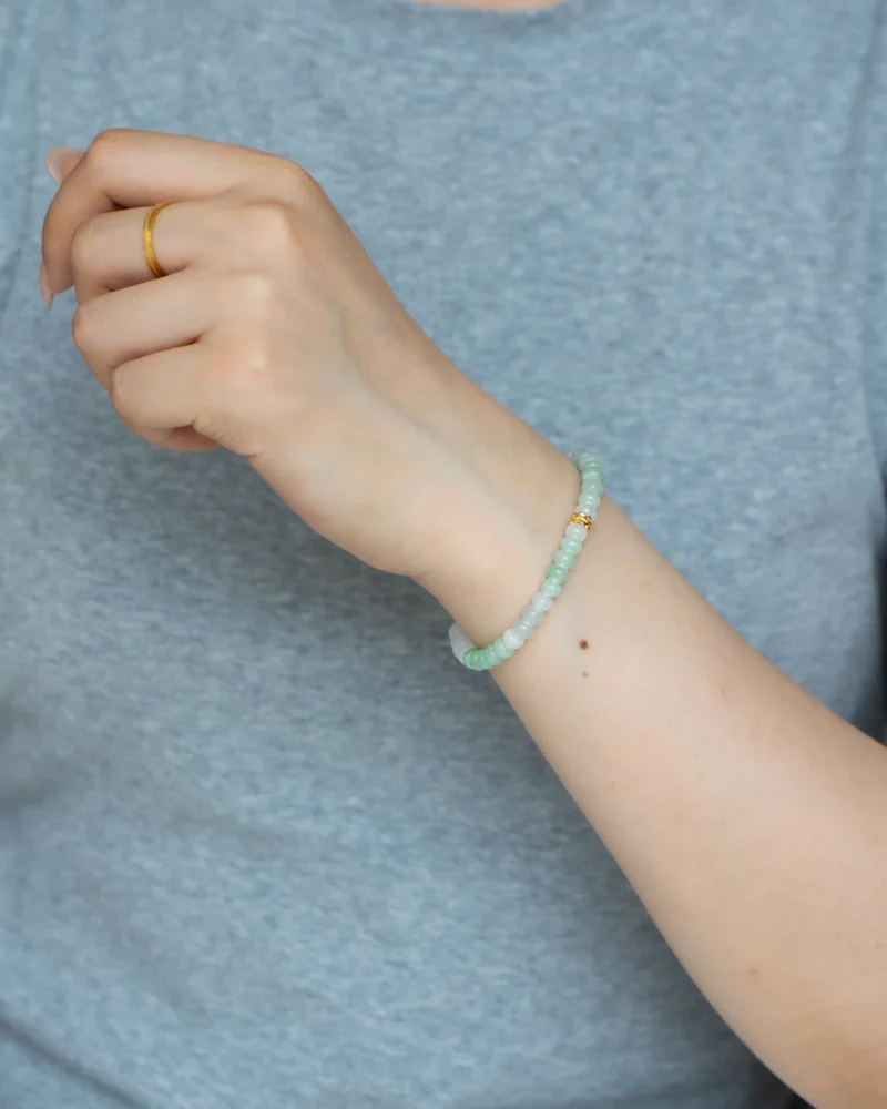 A person wearing a gold ring and the Brownish-Yellow, Light Green and White Jadeite Bracelet — featuring ring-shaped beads with gold-tone accent details — on their left hand, with a light gray shirt in the background.