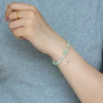 Someone wears a simple gold ring and a beaded green bracelet, paired with the Brownish-Yellow, Light Green and White Jadeite Bracelet—Ring-Shaped Beads with Gold-Tone Accent Details—on their left wrist, with a light gray shirt in the background.