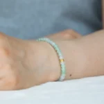 A close-up of a wrist wearing a Brownish-Yellow, Light Green and White Jadeite Bracelet with ring-shaped beads and gold-tone accent details, set against a soft, blurred background.