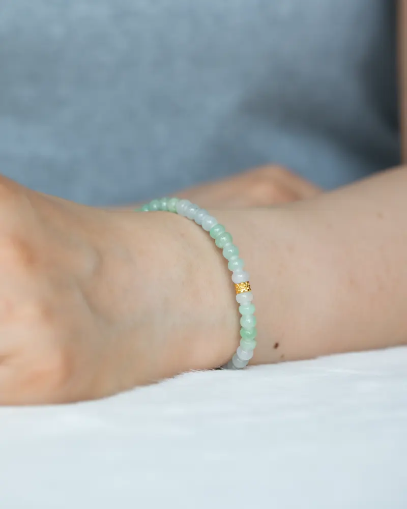 A close-up of a wrist wearing a Brownish-Yellow, Light Green and White Jadeite Bracelet with ring-shaped beads and gold-tone accent details, set against a soft, blurred background.