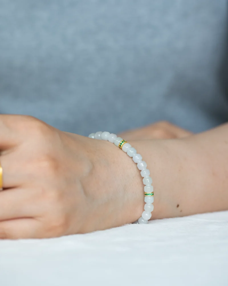 A close-up of a person’s wrist wearing the Light Green and White Jadeite Bracelet, featuring carved beads with gold-tone spacer details, against a light background.