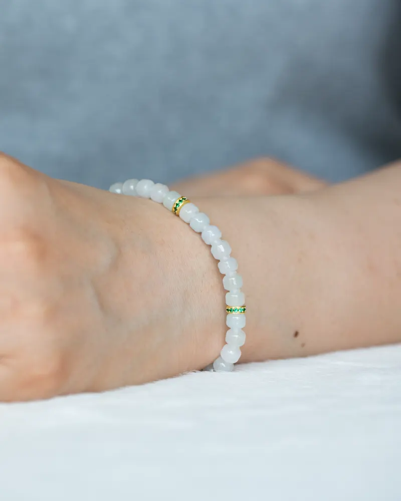 A close-up of a wrist wearing the Light Green and White Jadeite Bracelet, featuring carved beads with gold-tone spacer details. The background is blurred as the hand rests on a white surface.