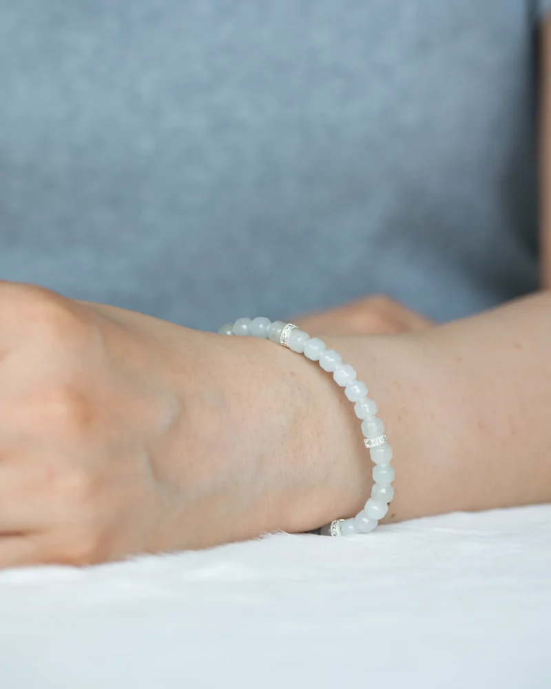 A person wearing the Light Green and White Jadeite Bracelet—featuring carved beads with silver-tone accent details—rests their arm on a white surface, dressed in a light gray shirt.