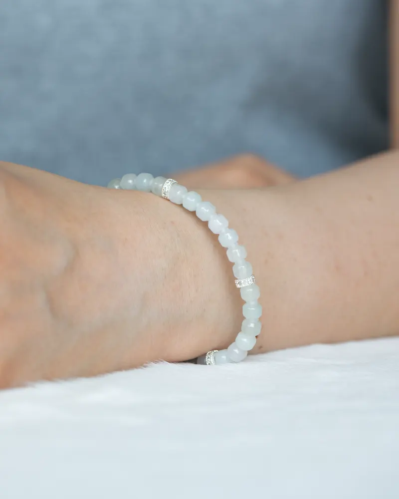 DSC_1187 拷贝 A person wears a light blue beaded bracelet with silver accents, wrist resting on a white surface. Nearby is the Light Green and White Jadeite Bracelet featuring carved beads and silver-tone accent details. The background is out of focus.