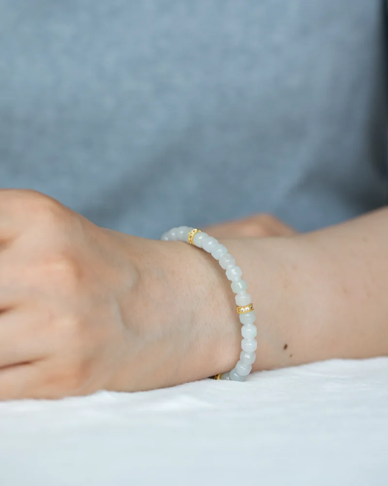 A person wearing a light grey beaded bracelet with small gold accents rests their arm on a white surface, against a soft grey background, complemented by a delicate Light Green and White Jadeite Bracelet featuring carved beads and gold-tone accent details.