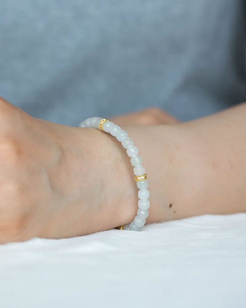 DSC_1188 拷贝 A person wears the Light Green and White Jadeite Bracelet—featuring carved beads with gold-tone accent details—on their wrist, set against a neutral background with their arm resting on a white surface.