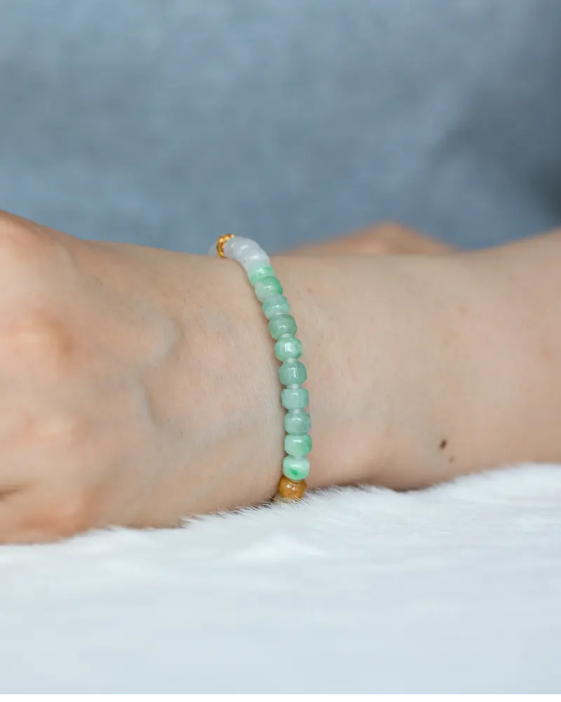 A close-up of a wrist wearing the Brownish-Yellow, Light Green and White Jadeite Bracelet featuring carved beads and gold-tone accent details, set against a soft, light background.