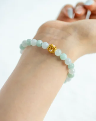 A light-skinned wrist wearing the Light Green and Greenish-White Jadeite Bracelet, featuring spherical beads and a gold-tone accent detail as the centerpiece, set against a soft white background.