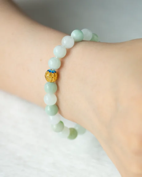 A Light Green and White Jadeite Bead Bracelet — Spherical Beads with a gold-tone accent is worn on a person’s wrist against a light background.