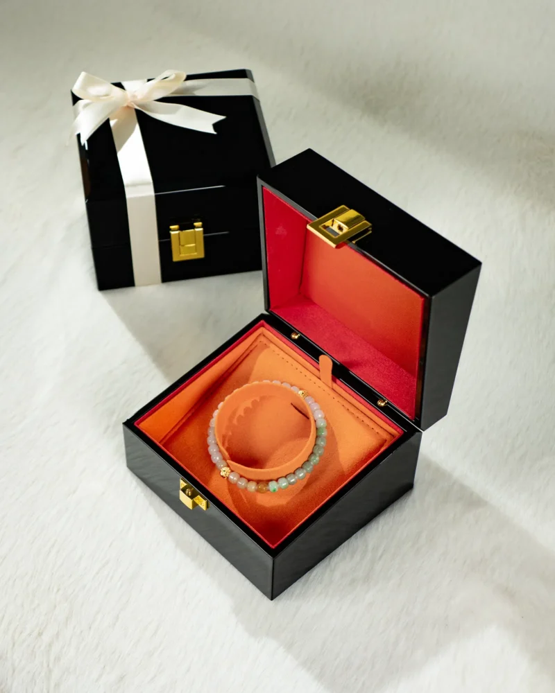 A Brownish-Yellow, Light Green and White Jadeite Bracelet — Carved Beads with Gold-Tone Accent Details rests in an open black jewelry box lined with orange fabric on a light surface. In the background, a closed black gift box tied with a white ribbon and finished with a gold clasp completes the presentation.