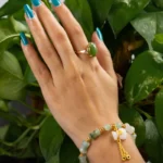 A hand with blue painted nails wears a green stone ring and a beaded bracelet with gold accents, posed against vibrant green leaves.