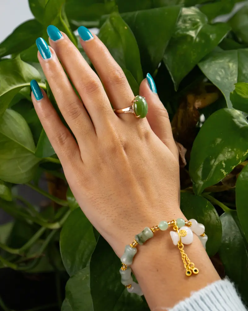 LYU_4175 拷贝 A hand with blue painted nails wears a green stone ring and a beaded bracelet with gold accents, posed against vibrant green leaves.