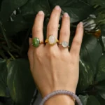 A hand wearing three gemstone rings and a lavender beaded bracelet is held against a background of green leafy plants.
