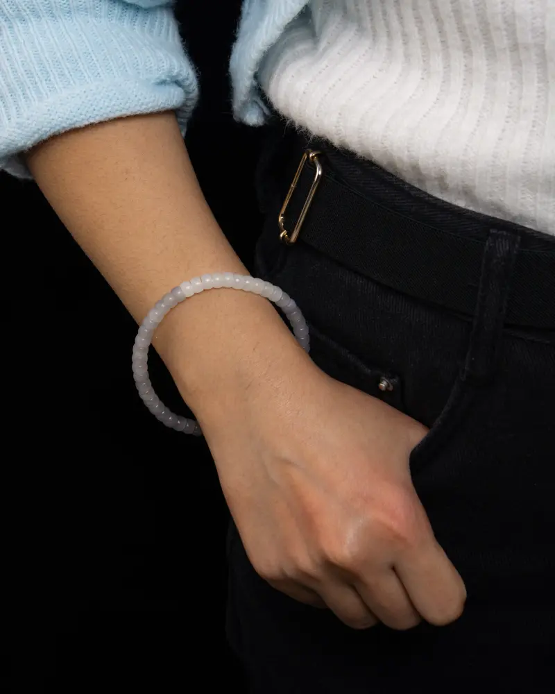 LYU_4253 拷贝 A person wearing a light blue sweater and white top has their hand in the pocket of dark pants, displaying a translucent beaded bracelet on their wrist against a black background.