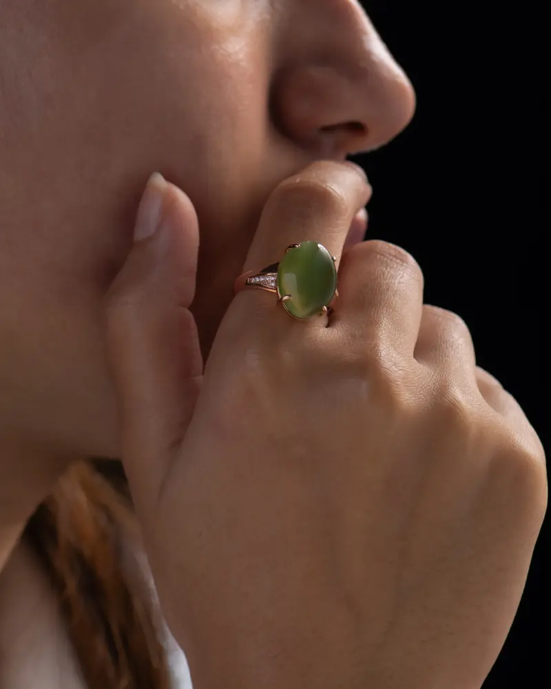LYU_4341 拷贝 A close-up of a person’s hand near their face, showcasing a large oval green gemstone ring set in a gold band against a dark background.