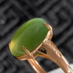 A close-up of a gold ring featuring an oval green gemstone and small accent diamonds along the band, set against a dark, textured background.