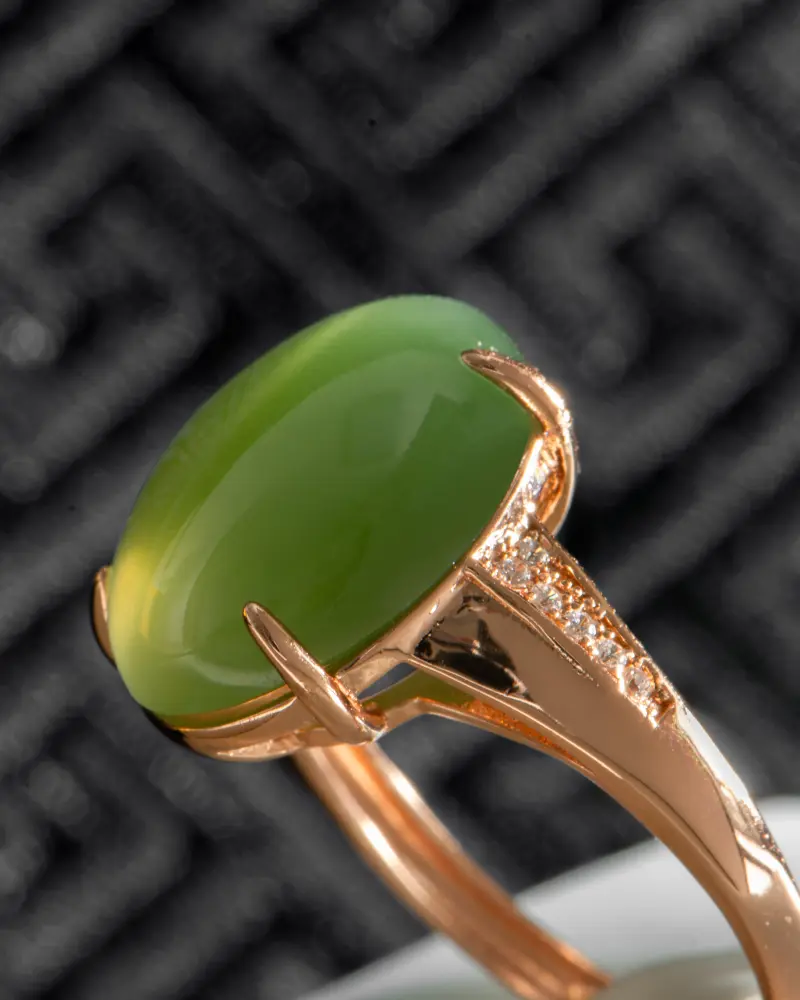 LYU_4477 拷贝 A close-up of a gold ring featuring an oval green gemstone and small accent diamonds along the band, set against a dark, textured background.