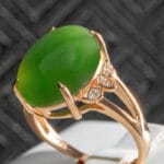 A gold ring with a large, oval green gemstone in the center, accented by three small round diamonds on one side. The ring is displayed against a dark textured background.