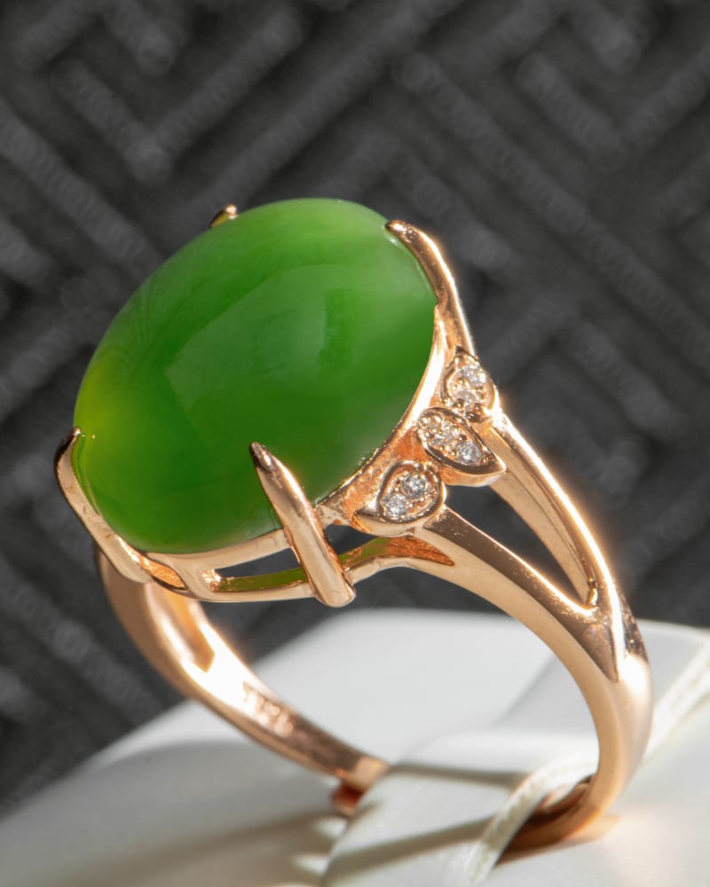A gold ring with a large, oval green gemstone in the center, accented by three small round diamonds on one side. The ring is displayed against a dark textured background.