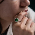 A close-up of a woman resting her chin on her hand, wearing a silver ring with a large green gemstone surrounded by small clear stones. The focus is on the ring and her thoughtful expression.