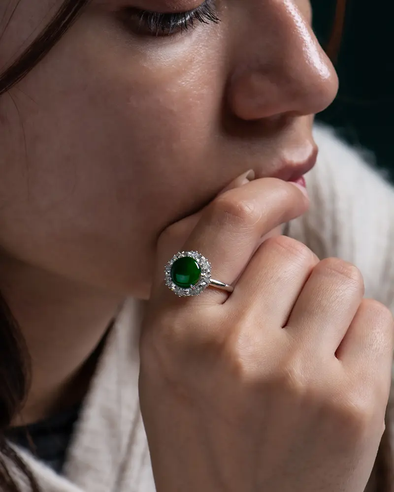 LYU_5149 拷贝 A close-up of a woman resting her chin on her hand, wearing a silver ring with a large green gemstone surrounded by small clear stones. The focus is on the ring and her thoughtful expression.