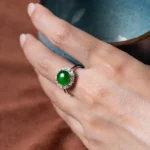A hand holding a blue ceramic cup, wearing a silver ring set with a large green gemstone surrounded by small clear stones. The background is a brown surface.