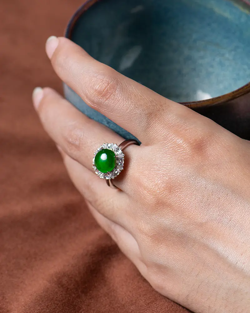 LYU_5158 拷贝 A hand holding a blue ceramic cup, wearing a silver ring set with a large green gemstone surrounded by small clear stones. The background is a brown surface.
