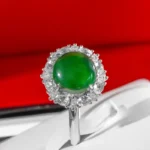 A silver ring featuring a round green gemstone surrounded by a halo of clear, sparkling stones, displayed on a white surface with a red background.