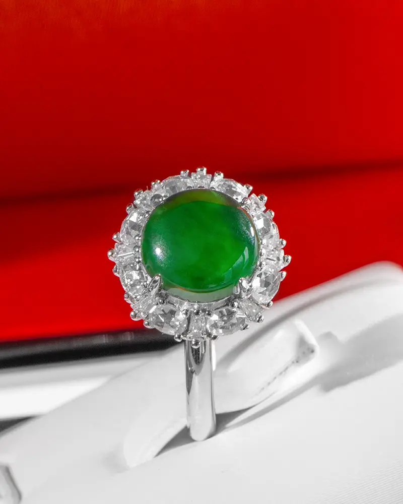 LYU_5234 拷贝 A silver ring featuring a round green gemstone surrounded by a halo of clear, sparkling stones, displayed on a white surface with a red background.