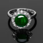 A silver ring featuring a large round green gemstone at the center, surrounded by a halo of clear, sparkling smaller stones, displayed on a reflective black surface.