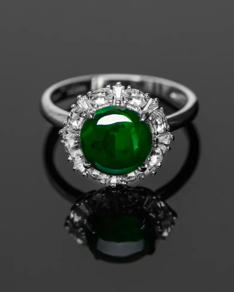 LYU_5246 拷贝 A silver ring featuring a large round green gemstone at the center, surrounded by a halo of clear, sparkling smaller stones, displayed on a reflective black surface.