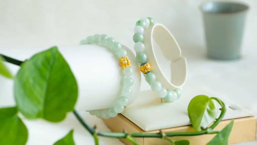Two jade bead bracelets with gold accents displayed on white stands, with green leaves and a soft-focus grey cup in the background.